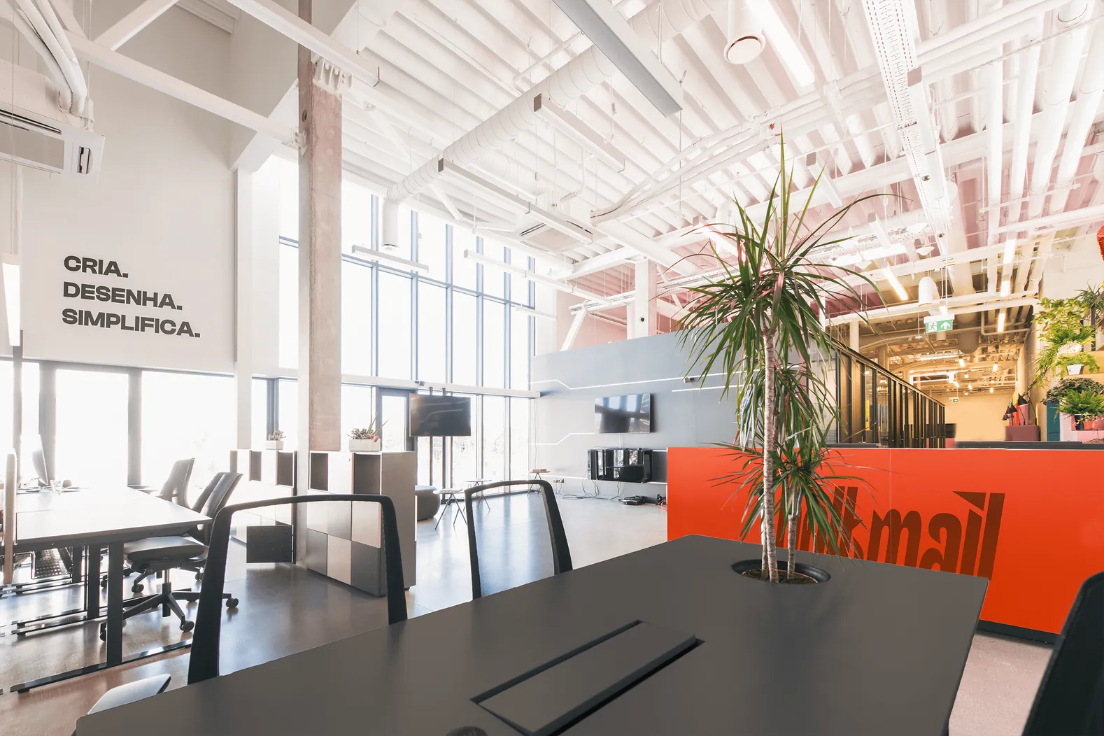 Open-plan Boltmail office with the line Cria. Desenha. Simplifica. on the wall and an orange reception desk
