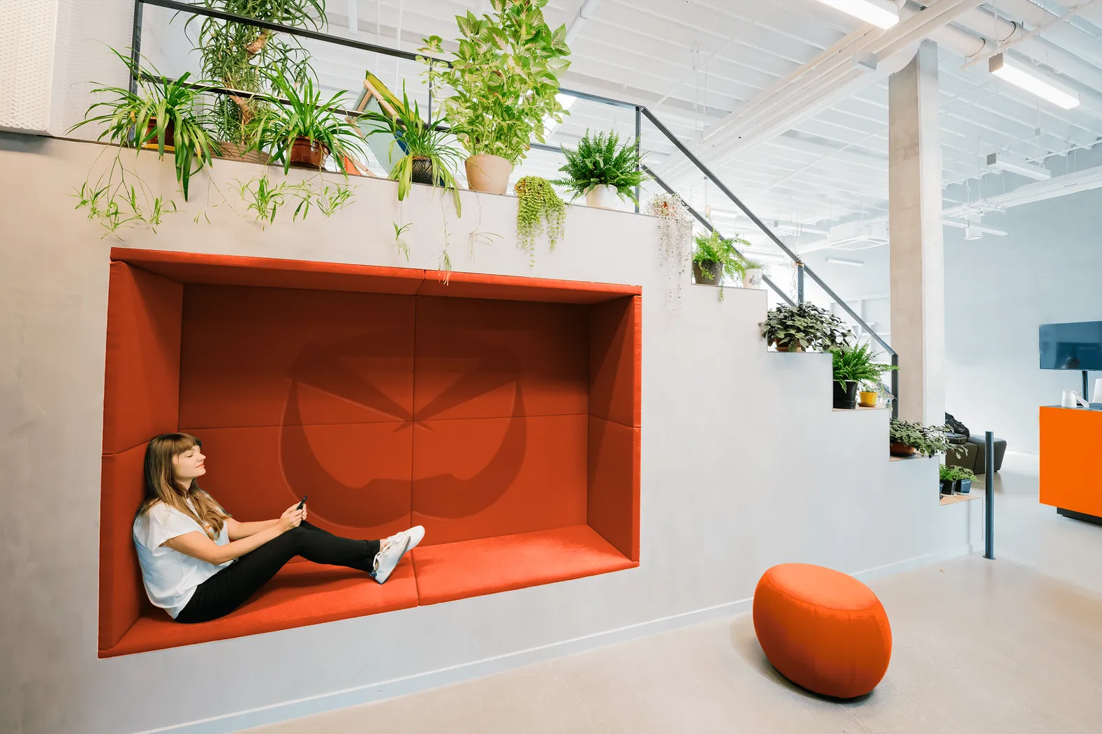 A reading nook in Boltmail orange, with the symbol pressed into the cushions, under a staircase of plants